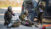 Mehrere Soldaten behandeln eine verletzte Person. Im Hintergrund sind zwei Militärfahrzeuge.