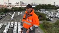 Ein Zivilist mit einer Warnjacke steht auf einer Erhöhung. Im Hintergrund sind ein Parkplatz und Kräne vom Hafen zu sehen.