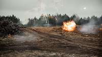 Eine freie Fläche mit Panzer und Explosionen, im Hintergrund Bäume