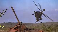 Ein Kampfhubschrauber fliegt schnell über ein braunes Panzerwrack auf einem bewaldeten Übungsplatz.