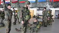 In einer Halle stehen und sitzen mehrere Soldaten mit ihrem Fallschirm, einer von ihnen wird gerade überprüft.