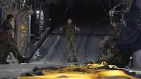 Ein Soldat steht im Innenraum des Transportflugzeuges. Links steht ein Soldat und an den Seiten sitzen drei Soldaten vor ihm.