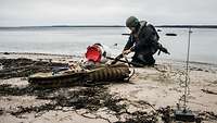 Ein Soldat in blauer Arbeitsuniform und mit Stahlhelm kniet an einem Strand vor einem tonnenförmigen Gegenstand.