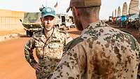 Ein Soldat mit blauem Basecap spricht mit einem anderen Soldaten. Im Hintergrund stehen Zelte und Fahrzeuge.