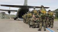 Soldaten mit Fallschirmgepäck stehen in zwei Reihen hinter einem Flugzeug. Ein Soldat überprüft einen Fallschirmspringer.