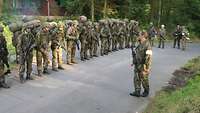 Mehrere Soldaten stehen mit mraschgepäck und Waffen in Reihe nebeneinander. Vor ihnen steht ein Ausbilder und spricht.