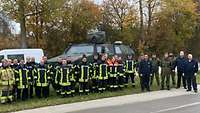 Mehrere Feuerwehrleute, Soldaten und zivile Mitarbeiter stehen am Rand einer Straße nebeneinander vor Fahrzeugen.