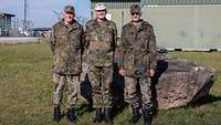Drei Männer in grüner Uniform stehen nebeneinander vor einem Stein auf einer grünen Wiese in einem Bundeswehr-Lager.