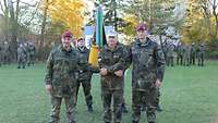 Drei Soldaten reichen sich auf einer Wiese die Hände. Hinter ihnen stehen eine Fahnenabordnung und Soldaten in Formation.