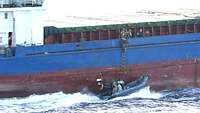 Dicht an einem Schiff fährt ein Speedboot. An der Bordwand des Schiffes klettern Soldaten an einer Strickleiter hoch.