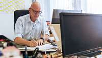 Mann mit Brille und weiß-blauem Hemd, sitzt an seinem Arbeitsplatz vor seinem Computer