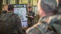 Ein Soldat steht im Wald vor einer aufgestellten Lagekarte und spricht. Soldaten stehen drumherum.
