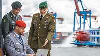 Drei Soldaten blicken interessiert auf ein Notebook, im Hintergrund ist der Hamburger Hafen zu sehen