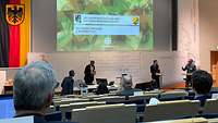 Blick aus den Zuschauerrängen auf das Podium des Symposiums, auf dem sich der zivile Moderator sowie zwei Soldaten befinden.