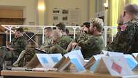 Mehrere Soldaten hören einem Vortrag zu. Im Vordergrund stehen auf einem Tisch aus Pappe gebaute militärische Symbole.