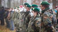 Soldaten in Flecktarnuniform und Zivilisten stehen in einer seitlichen Reihe.