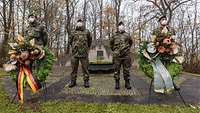 Vier Soldaten stehen vor einem Ehrenmal. Vor ihnen sind zwei Kränze aufgestellt.