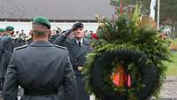 Zwei Soldaten stehen neben einem Kranz, ein Soldat grüßt militärisch. Im Hintergrund spielt ein Musikkorps.