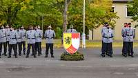 Ein Wappenschild in der Bildmitte, links und rechts daneben angetretene Soldaten.