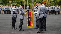 Soldaten mit einer schwarz-rot-goldenen Flagge, ein General befestigt daran ein gelbes Fahnenband.