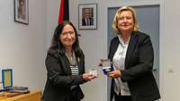 Zwei weibliche Personen in einem Dienstzimmer präsentieren jeweils eine Medaille.