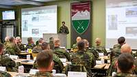 In einem Saal sitzen Soldaten in Reihen an Tischen. Vor ihnen steht ein Soldat vor einer Projektionswand am Rednerpult.