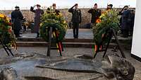 Eine Gruppe von Soldaten steht vor einem Ehrenmal mit Blumenkränzen und grüßt militärisch. 