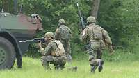 Ein Soldat kniet neben einem gepanzerten Fahrzeug und sichert seine Kameraden. Zwei weitere Soldaten laufen hinter ihm vorbei.