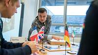 Ein Soldat steht am Tisch mit einem aufgebauten Brettspiel und erklärt einem Mann etwas.
