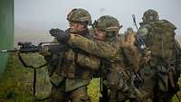 Eine Gruppe Soldaten versteckt sich im Gras bei Nebel hinter einer Hauswand und gibt sich Handzeichen.