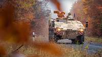 Ein beigefarbenes Gefechtsfahrzeug fährt auf einem Schotterweg durch den herbstlichen Laubwald.
