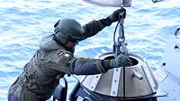 Ein Soldat hält einen großen Metallhaken in den Händen, an dem ein Boot hängt 