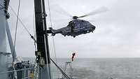 Ein mittelgroßer, blauer Hubschrauber schwebt über dem Heck eines kleinen Schiffes in See.