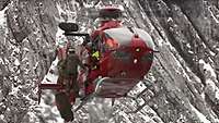 Vor einer grau-weißen schroffen Gebirgswand schwebt ein roter Hubschrauber, vor ihm hängen Menschen an Seilen.