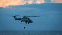 Ein grauer Hubschrauber fliegt über See, unter ihm hängt ein angebrachtes Sonar.