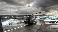 Ein grauer Hubschrauber auf dem Flugdeck eines grauen Schiffes bei schwerer See.