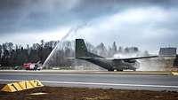 Die Wasserfontäne eines Feuerwehrfahrzeugs geht auf ein Militärflugzeug nieder. Beide stehen auf einer Landebahn.