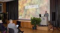 Ein Soldat an einem Rednerpult vor einer Leinwand mit dem Wort Vernissage