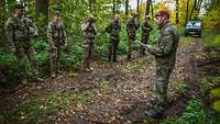 Ein deutscher Soldat mit einem Zettel in der Hand steht vor Soldaten verschiedener Nationen auf einem Waldweg und spricht.
