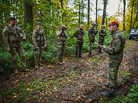 Ein deutscher Soldat mit einem Zettel in der Hand steht vor Soldaten verschiedener Nationen auf einem Waldweg und spricht.