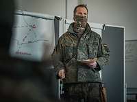 Ein Soldat hält in einem weißen Raum vor einer grauen Tafel einen Vortrag.