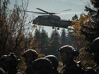 Hinter einer Gruppe Soldaten landet auf einer Waldlichtung ein großer dunkler Hubschrauber.
