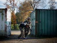 Ein Soldat rennt mit einer festgenommenen Person zwischen wie Containern über eine Sandfläche.
