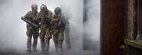 Drei Soldaten laufen mit ihrem Gewehr bei Rauch an einem Gebäude vorbei.