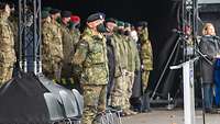 Ein Soldat mit schwarzem Mund-Nasenschutz steht seitlich von einer Tribüne mit Gästen.