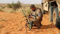 Ein Soldat kniet im Sand hinter einem Funkgerät und bedient es