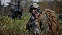 Ein Soldat sichert das Gelände mit seiner Waffe auf einer Wiese, im Hintergrund startet ein Hubschrauber.