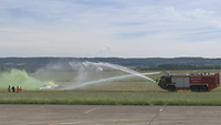 Mit einem Wasser-Doppelstrahl geht das Feuerwehrauto gegen gelben Rauch vor. In der Nähe des Rauchs sind Menschen.