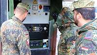 Drei Soldaten stehen vor einer Maschine, die bei der Dekontamination von Personal und Material unterstützt. 