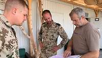 Zwei Soldaten stehen einem Zivilisten gegenüber. Der Zivilist hält mehrere Papiere in der Hand.